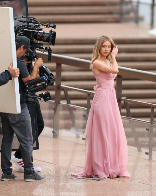 Sydney Sweeney with Glen Powell filming scenes for their upcoming untitled film in Sydney, Australia - March 27 2023

Sydney Sweeney Embraces Rom-Com Dressing in Flowy Pink Dress & Sparkling Sandals While Filming With Glen Powell in Australia

Sydney Sweeney was seen on Monday at the steps of the Sydney Opera House in Sydney, filming for her unnamed upcoming rom-com alongside leading man Glen Powell.

Sweeney and Powell were seen on the steps of the well-known performance center in rom-com-ready ensembles. Powell, who has previously starred in romantic features like “Set It Up,” wore a classic black-and-white tuxedo with black leather point-toe oxfords.

Whereas Sweeney looked every bit the leading lady in a flowing, deeply-cut, deftly-wrapped pink gown with a gauzy overlay of fabric over the skirt. The actress wore her well-known blond locks in a center part and styled them into soft, romantic waves.

For shoes, Sweeney donned a pair of peep-toe silver stiletto heels. The style featured a glittery finish and a ankle-strap for extra security.

When she’s not on set, Sweeney can be seen rocking a variety of footwear styles from ladylike mules to sky-high stilettos. She has been seen wearing a variety of designers, from Jimmy Choo to Tory Burch. Sweeney has been a Tory Burch brand ambassador since Apr. 2022, notably starring in the designer’s summer 2022 campaign, highlighting her popular Miller sandal line.

Sweeney has gained worldwide recognition after playing the love-obsessed Cassie on the hit HBO series “Euphoria”, and in the past year has been stepping into a variety of roles outside of the dramatic high school archetype.

On Jan. 11, Deadline reported that Sweeney and Powell signed on to star in a to-be-named rated-R romantic comedy film directed by Will Gluck. Not only is Sweeney the leading lady of the film, but she is also serving as the project’s executive producer.