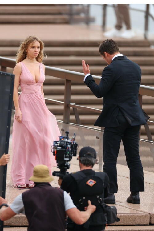 Sydney Sweeney with Glen Powell filming scenes for their upcoming untitled film in Sydney, Australia - March 27 2023

Sydney Sweeney Embraces Rom-Com Dressing in Flowy Pink Dress & Sparkling Sandals While Filming With Glen Powell in Australia

Sydney Sweeney was seen on Monday at the steps of the Sydney Opera House in Sydney, filming for her unnamed upcoming rom-com alongside leading man Glen Powell.

Sweeney and Powell were seen on the steps of the well-known performance center in rom-com-ready ensembles. Powell, who has previously starred in romantic features like “Set It Up,” wore a classic black-and-white tuxedo with black leather point-toe oxfords.

Whereas Sweeney looked every bit the leading lady in a flowing, deeply-cut, deftly-wrapped pink gown with a gauzy overlay of fabric over the skirt. The actress wore her well-known blond locks in a center part and styled them into soft, romantic waves.

For shoes, Sweeney donned a pair of peep-toe silver stiletto heels. The style featured a glittery finish and a ankle-strap for extra security.

When she’s not on set, Sweeney can be seen rocking a variety of footwear styles from ladylike mules to sky-high stilettos. She has been seen wearing a variety of designers, from Jimmy Choo to Tory Burch. Sweeney has been a Tory Burch brand ambassador since Apr. 2022, notably starring in the designer’s summer 2022 campaign, highlighting her popular Miller sandal line.

Sweeney has gained worldwide recognition after playing the love-obsessed Cassie on the hit HBO series “Euphoria”, and in the past year has been stepping into a variety of roles outside of the dramatic high school archetype.

On Jan. 11, Deadline reported that Sweeney and Powell signed on to star in a to-be-named rated-R romantic comedy film directed by Will Gluck. Not only is Sweeney the leading lady of the film, but she is also serving as the project’s executive producer.