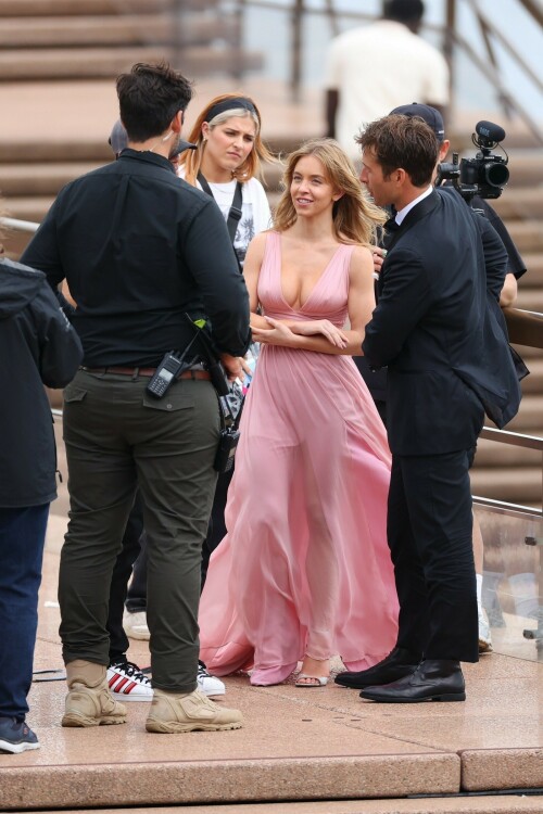 Sydney Sweeney with Glen Powell filming scenes for their upcoming untitled film in Sydney, Australia - March 27 2023

Sydney Sweeney Embraces Rom-Com Dressing in Flowy Pink Dress & Sparkling Sandals While Filming With Glen Powell in Australia

Sydney Sweeney was seen on Monday at the steps of the Sydney Opera House in Sydney, filming for her unnamed upcoming rom-com alongside leading man Glen Powell.

Sweeney and Powell were seen on the steps of the well-known performance center in rom-com-ready ensembles. Powell, who has previously starred in romantic features like “Set It Up,” wore a classic black-and-white tuxedo with black leather point-toe oxfords.

Whereas Sweeney looked every bit the leading lady in a flowing, deeply-cut, deftly-wrapped pink gown with a gauzy overlay of fabric over the skirt. The actress wore her well-known blond locks in a center part and styled them into soft, romantic waves.

For shoes, Sweeney donned a pair of peep-toe silver stiletto heels. The style featured a glittery finish and a ankle-strap for extra security.

When she’s not on set, Sweeney can be seen rocking a variety of footwear styles from ladylike mules to sky-high stilettos. She has been seen wearing a variety of designers, from Jimmy Choo to Tory Burch. Sweeney has been a Tory Burch brand ambassador since Apr. 2022, notably starring in the designer’s summer 2022 campaign, highlighting her popular Miller sandal line.

Sweeney has gained worldwide recognition after playing the love-obsessed Cassie on the hit HBO series “Euphoria”, and in the past year has been stepping into a variety of roles outside of the dramatic high school archetype.

On Jan. 11, Deadline reported that Sweeney and Powell signed on to star in a to-be-named rated-R romantic comedy film directed by Will Gluck. Not only is Sweeney the leading lady of the film, but she is also serving as the project’s executive producer.