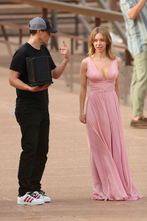 Sydney Sweeney with Glen Powell filming scenes for their upcoming untitled film in Sydney, Australia - March 27 2023

Sydney Sweeney Embraces Rom-Com Dressing in Flowy Pink Dress & Sparkling Sandals While Filming With Glen Powell in Australia

Sydney Sweeney was seen on Monday at the steps of the Sydney Opera House in Sydney, filming for her unnamed upcoming rom-com alongside leading man Glen Powell.

Sweeney and Powell were seen on the steps of the well-known performance center in rom-com-ready ensembles. Powell, who has previously starred in romantic features like “Set It Up,” wore a classic black-and-white tuxedo with black leather point-toe oxfords.

Whereas Sweeney looked every bit the leading lady in a flowing, deeply-cut, deftly-wrapped pink gown with a gauzy overlay of fabric over the skirt. The actress wore her well-known blond locks in a center part and styled them into soft, romantic waves.

For shoes, Sweeney donned a pair of peep-toe silver stiletto heels. The style featured a glittery finish and a ankle-strap for extra security.

When she’s not on set, Sweeney can be seen rocking a variety of footwear styles from ladylike mules to sky-high stilettos. She has been seen wearing a variety of designers, from Jimmy Choo to Tory Burch. Sweeney has been a Tory Burch brand ambassador since Apr. 2022, notably starring in the designer’s summer 2022 campaign, highlighting her popular Miller sandal line.

Sweeney has gained worldwide recognition after playing the love-obsessed Cassie on the hit HBO series “Euphoria”, and in the past year has been stepping into a variety of roles outside of the dramatic high school archetype.

On Jan. 11, Deadline reported that Sweeney and Powell signed on to star in a to-be-named rated-R romantic comedy film directed by Will Gluck. Not only is Sweeney the leading lady of the film, but she is also serving as the project’s executive producer.