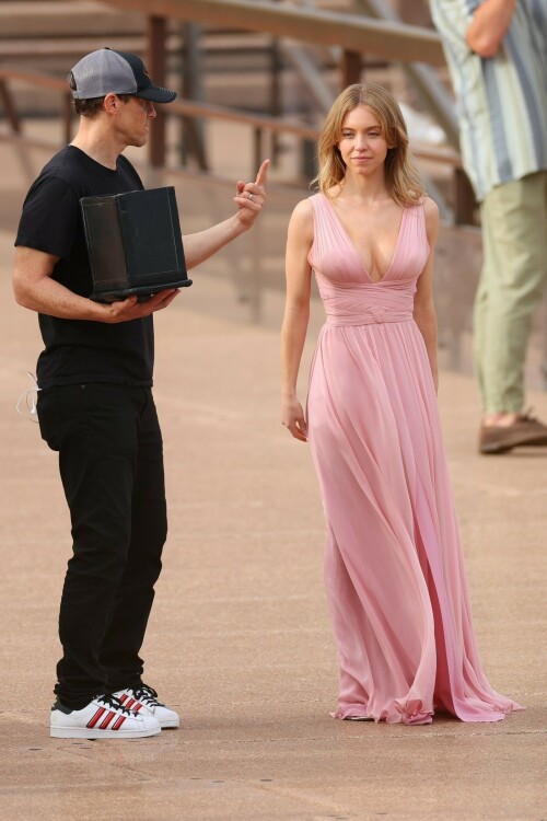 Sydney Sweeney with Glen Powell filming scenes for their upcoming untitled film in Sydney, Australia - March 27 2023

Sydney Sweeney Embraces Rom-Com Dressing in Flowy Pink Dress & Sparkling Sandals While Filming With Glen Powell in Australia

Sydney Sweeney was seen on Monday at the steps of the Sydney Opera House in Sydney, filming for her unnamed upcoming rom-com alongside leading man Glen Powell.

Sweeney and Powell were seen on the steps of the well-known performance center in rom-com-ready ensembles. Powell, who has previously starred in romantic features like “Set It Up,” wore a classic black-and-white tuxedo with black leather point-toe oxfords.

Whereas Sweeney looked every bit the leading lady in a flowing, deeply-cut, deftly-wrapped pink gown with a gauzy overlay of fabric over the skirt. The actress wore her well-known blond locks in a center part and styled them into soft, romantic waves.

For shoes, Sweeney donned a pair of peep-toe silver stiletto heels. The style featured a glittery finish and a ankle-strap for extra security.

When she’s not on set, Sweeney can be seen rocking a variety of footwear styles from ladylike mules to sky-high stilettos. She has been seen wearing a variety of designers, from Jimmy Choo to Tory Burch. Sweeney has been a Tory Burch brand ambassador since Apr. 2022, notably starring in the designer’s summer 2022 campaign, highlighting her popular Miller sandal line.

Sweeney has gained worldwide recognition after playing the love-obsessed Cassie on the hit HBO series “Euphoria”, and in the past year has been stepping into a variety of roles outside of the dramatic high school archetype.

On Jan. 11, Deadline reported that Sweeney and Powell signed on to star in a to-be-named rated-R romantic comedy film directed by Will Gluck. Not only is Sweeney the leading lady of the film, but she is also serving as the project’s executive producer.